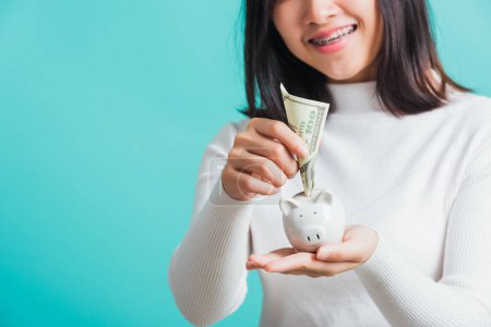 Photo for Asian beautiful young woman holding piggy bank on hands and putting dollars money banknotes, portrait of happy female smiling hold piggybank in palm, isolated on blue background, saving money concept - Royalty Free Image