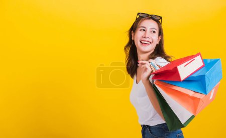 Foto de Asian Thai portrait happy beautiful cute young woman smiling stand with sunglasses excited holding shopping bags multi color looking back, studio shot isolated yellow background with copy space - Imagen libre de derechos