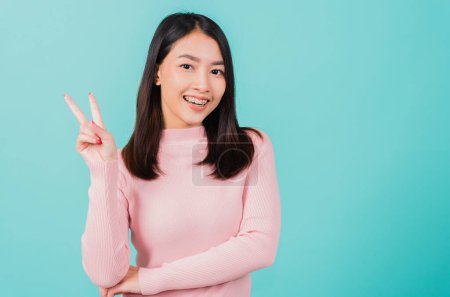 Téléchargez les photos : Jeune asiatique belle femme souriante porter des attaches orthodontiques en silicone sur les dents montrant doigt de victoire de signe V isolé sur fond bleu, en conservant après bretelles amovibles. Concept d'hygiène dentaire - en image libre de droit