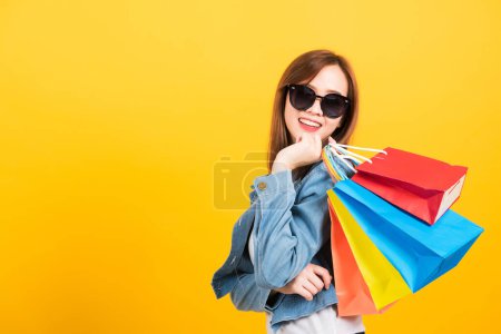 Photo for Asian happy portrait beautiful cute young woman teen smiling standing with sunglasses excited holding shopping bags multi color looking camera isolated, studio shot yellow background with copy space - Royalty Free Image