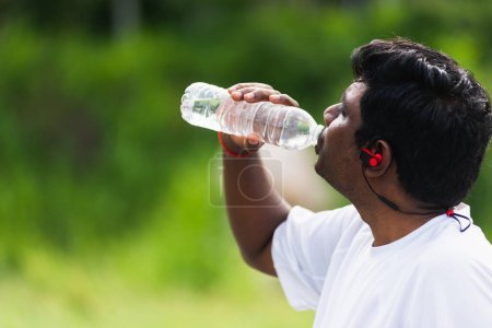 Téléchargez les photos : Gros plan Asiatique jeune coureur de sport homme noir porter des écouteurs d'athlète il boire de l'eau d'une bouteille après avoir couru au parc de santé de la rue en plein air, concept d'entraînement d'exercice sain - en image libre de droit