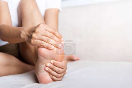 Photo for Foot pain, Asian woman sitting on sofa feeling pain in her foot at home, female suffering from feet ache use hand massage relax muscle from soles in home interior, Healthcare and podiatry medical - Royalty Free Image