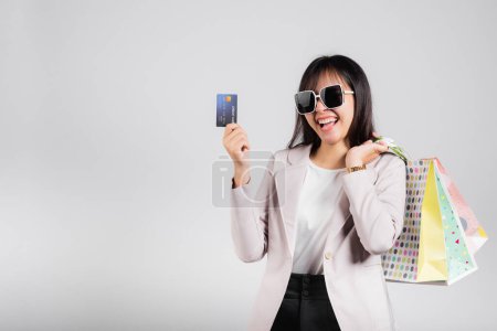 Téléchargez les photos : Femme avec des lunettes sourire client confiant tenir sacs à provisions en ligne et carte de crédit pour le paiement à portée de main, Portrait heureux asiatique heureux magasin de mode femme studio tourné isolé sur fond blanc - en image libre de droit