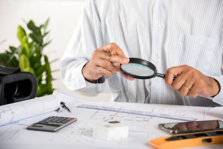 Foto de Architectural interior designer working holding magnifying glass and look on blueprint in working site paper project plans on desk table, engineer tool contractor building, Architect workplace concept - Imagen libre de derechos