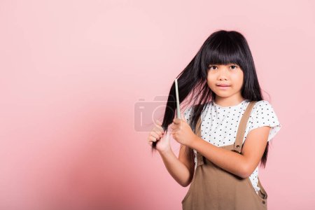 Téléchargez les photos : Asiatique petit enfant 10 ans tenir peigne brossage son indiscipliné elle toucher ses longs cheveux noirs au studio tourné isolé sur fond rose, Happy child girl avec une brosse à cheveux, concept de soins des cheveux - en image libre de droit