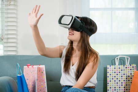 Foto de Happy woman customer with shopping bags around wear VR headset on sofa touching air, Asian young female wearing modern virtual reality headsets have experience shopping online in living room at home - Imagen libre de derechos