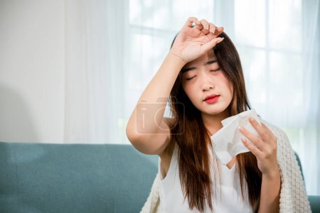 Foto de Asian young woman she sick blowing nose sneezing in tissue at home, fever caught cold, Sick female sitting under blanket on sofa and sneeze with tissue paper in living room, medicine healthcare - Imagen libre de derechos