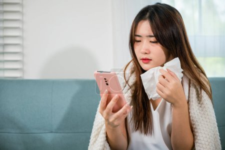 Téléchargez les photos : Asian young woman sick sitting down on sofa with cold blowing her nose and checking smart mobile phone to cell to doctor online, depressed sad sick female holding tissues and smartphone in living room - en image libre de droit