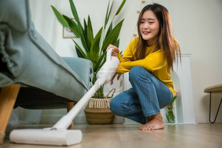 Foto de Happy Asian young woman with accumulator vacuum cleaner at home in living room, housewife female dust cleaning floor under sofa or furniture with vacuum cleaner, household and housework concept - Imagen libre de derechos