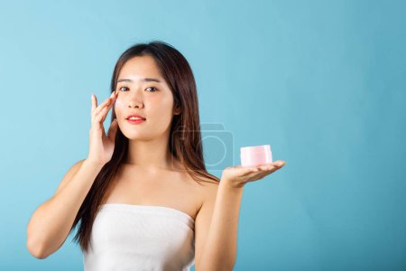 Photo for Asian young woman are not very beautiful hold jar of skin cream for face and apply anti-aging moisturizer studio isolated on blue background, Happy female pointing finger her face, pursuit of beauty - Royalty Free Image