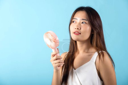 Asian beauty young woman holding pink portable electric mini fan near her face studio shot isolated on blue background, Female hand hold small plastic fan handheld she enjoying cool wind blowing