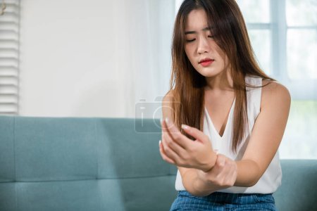 Photo for Closeup hand. Asian young woman sitting on sofa holds suffering her wrist at home, female hand injury feeling joint pain, performing self-massage, Health care and medical concept - Royalty Free Image