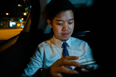 Foto de Handsome Asian young man in taxi work on late night sitting back seat and still work with mobile phone in city street, Businessman using smartphone in backseat of car near window at night - Imagen libre de derechos