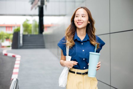 Photo for Asian beautiful business woman confident smiling with cloth bag holding steel thermos tumbler mug water glass she walking outdoors on street near modern building office, Happy female looking side away - Royalty Free Image