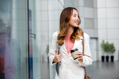 Photo for Young business woman wearing white suit jacket and smile while go to work outdoor mirror building background. Confident Businesswoman with cup of coffee holding Laptop walking outside office building - Royalty Free Image