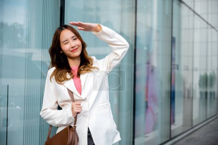 Photo for Happy woman wearing white suit jacket with brown bag at sunlight outdoors. Portrait of confidence young businesswoman standing outside office building in city raise your hand to shade sun - Royalty Free Image