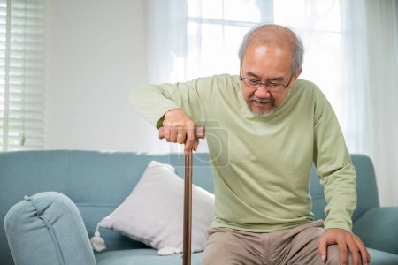 Photo for Asian senior old man with eyeglasses type to standing up from sofa with walking cane stick to walk at home, Elderly suffering from knee pain ache holding handle of cane, retirement medical healthcare - Royalty Free Image