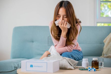 Foto de Sick woman. Asian young girl cold sick she sneeze with tissue paper on sofa, beautiful female health problem blowing nose use pharmacy kit box delivery service from hospital, delivery pharmacy concept - Imagen libre de derechos