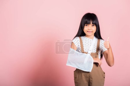 Photo for Arm broken. Asian little kid 10 years old hand bone broken from accident with arm splint show thumb up finger at studio shot isolated on pink background, Happy child girl accident insurance - Royalty Free Image