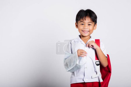 Photo for Asian student kid boy wearing student thai uniform accident broken bone wearing splint arm plaster fiberglass cast covering arm in cast at studio shot isolated on white background, back to school - Royalty Free Image