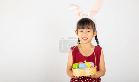 Foto de Feliz día de Pascua. Sonríe niña asiática con orejas de conejito de Pascua sosteniendo cesta de huevos llenos de colores sonrisas ampliamente aislados en el fondo blanco con espacio para copiar, niño feliz en vacaciones - Imagen libre de derechos