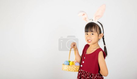 Téléchargez les photos : Joyeuse fête de Pâques. Sourire petite fille asiatique portant des oreilles de lapin de Pâques tenant panier d'oeufs pleins de couleurs sourit largement isolé sur fond blanc avec espace de copie, Joyeux enfant en vacances - en image libre de droit