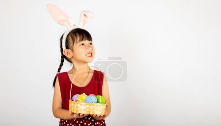 Foto de Feliz día de Pascua. Sonríe niña asiática con orejas de conejito de Pascua sosteniendo cesta de huevos llenos de colores sonrisas ampliamente aislados en el fondo blanco con espacio para copiar, niño feliz en vacaciones - Imagen libre de derechos