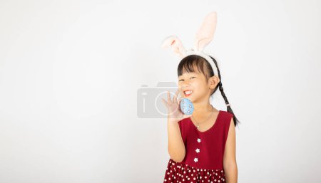 Téléchargez les photos : Joyeuse fête de Pâques. Sourire petite fille asiatique portant des oreilles de lapin de Pâques tenant des œufs colorés sur des mains isolées sur fond blanc avec espace de copie, Joyeux enfant en vacances - en image libre de droit