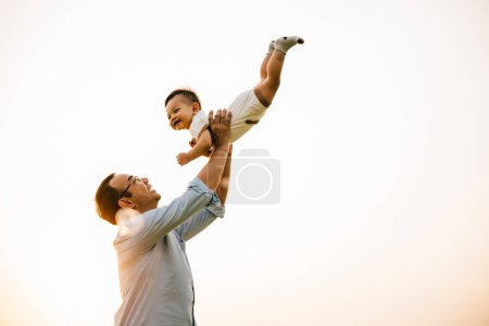 Photo for In a beautiful moment of family togetherness, a father holds his baby daughter up high, throwing her up into the sunny sky at sunset. The cheerful child smiles with freedom and playful fun - Royalty Free Image