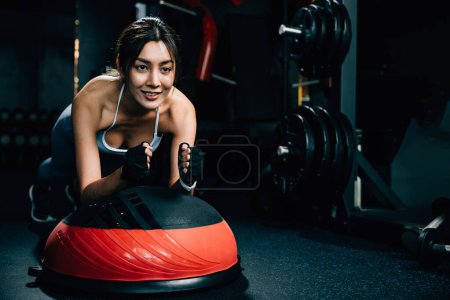 Foto de Mujer asiática delgada realizando un ejercicio de tablón en una pelota de estabilidad, demostrando equilibrio y agilidad mientras mejora su forma corporal general, ejercicio en el fondo oscuro del gimnasio - Imagen libre de derechos