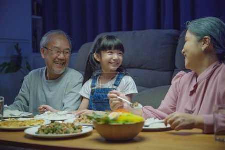 Foto de Feliz familia asiática abuela abuelo y nieta cenar en la mesa y divertirse durante la noche en casa, padre mayor y el niño comer juntos en la sala de estar en interiores juntos - Imagen libre de derechos