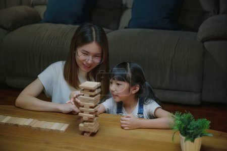 Téléchargez les photos : Femme souriante aider à enseigner le jeu d'enfant construire constructeur de blocs de bois, Asiatique jeune mère jouer à un jeu en bloc de bois avec sa petite fille dans le salon à la maison la nuit avant d'aller au lit - en image libre de droit