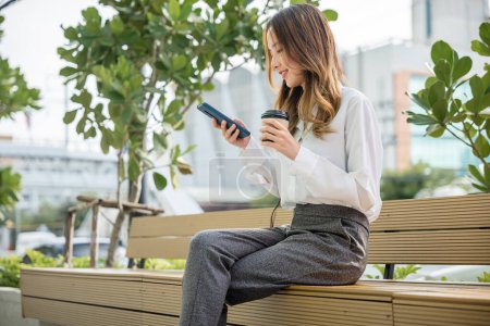 Photo for Smiling woman sitting on bench outdoor on the park using smartphone chatting social media, Happy Asian businesswoman using mobile phone, technology communication, coffee paper cup takeaway - Royalty Free Image