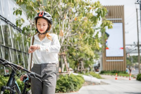 Téléchargez les photos : Asiatique jeune femme d'affaires debout avec vélo vérifier l'heure sur la rue de la ville le matin avant d'aller au travail au bureau, portrait souriant femme vérifier montre, Trajet vers le travail, mode de vie motard - en image libre de droit