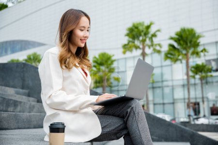 Téléchargez les photos : Heureuse jeune femme asiatique freelance regarder ordinateur portable avec le sourire tout en travaillant avec tasse de café tout en étant assis sur les escaliers à l'extérieur dans la ville. Concept de travail à distance dans le bureau. - en image libre de droit