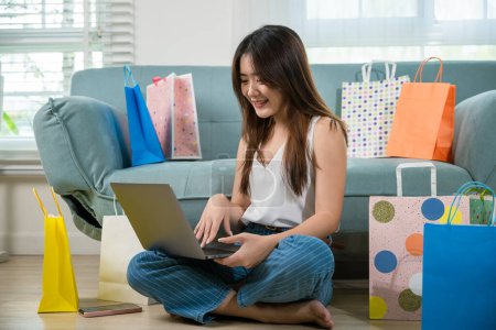 Foto de Mujer joven asiática sentada en un sofá con bolsas de compras usando computadora portátil para ir de compras en línea, feliz cliente satisfecho con la compra en el sitio web en la sala de estar en casa - Imagen libre de derechos