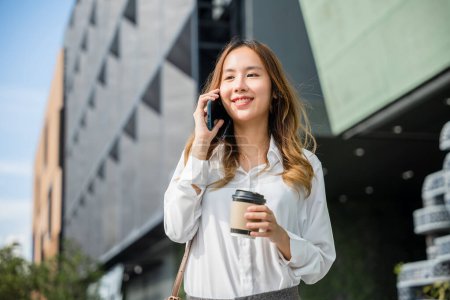 Photo for Asian young businesswoman walking outdoors talking on cell smart mobile phone and holding coffee cup takeaway, lifestyle business woman hold paper cup and calling smartphone outside office - Royalty Free Image