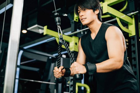 Foto de Hombre asiático usando una máquina de cable para hacer tiradas de tríceps, centrándose en la construcción de la fuerza y el mantenimiento de un estilo de vida saludable. Ejercicio de fuerza superior - Imagen libre de derechos