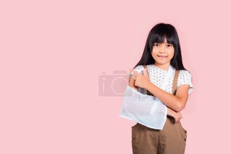 Photo for Arm broken. Asian little kid 10 years old hand bone broken from accident with arm splint pointing finger to copy space at studio shot isolated on pink background, Happy child girl accident insurance - Royalty Free Image