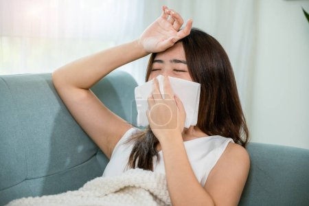 Foto de Mujer enferma sentada debajo de la manta en el sofá y estornudar con papel de seda en la sala de estar, Asiática joven hembra que sopla la nariz estornudos en el tejido en casa, la fiebre se resfrió - Imagen libre de derechos