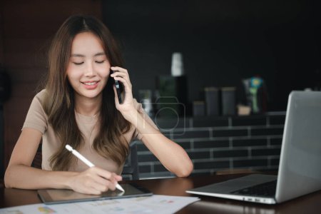 Photo for Asian businesswoman sitting at cafe calling mobile phone talking with customer asking about something, smiling female working laptop computer and writing take note on digital tablet at home office - Royalty Free Image