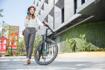 Foto de Asiática sonriente mujer joven con casco celebrar teléfono inteligente móvil hablar con los negocios durante ir al trabajo de oficina en la calle con la bicicleta, respetuoso del medio ambiente, estilo de vida negocio femenino conmutar al aire libre en la mañana - Imagen libre de derechos