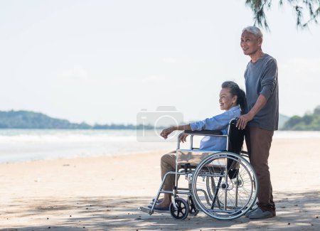 Téléchargez les photos : Happy Asian dos femme âgée handicapé assis en fauteuil roulant et le mari est un utilisateur de fauteuil roulant sur la plage ensemble, vacances d'été, concept de couple de retraite - en image libre de droit