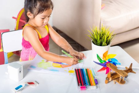 Téléchargez les photos : Enfant heureux petite fille coloré paysage de dessin mon rêve à la maison sur papier, Asiatique enfant mignon préscolaire assis sur la table sourire elle dessine image maison de campagne avec crayon à la maison pour apprendre les arts devoirs - en image libre de droit