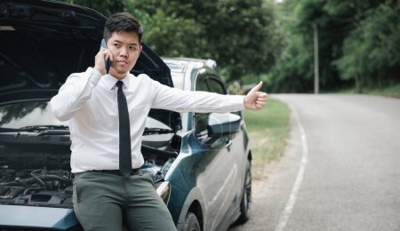 Téléchargez les photos : Homme d'affaires asiatique échoué sur le bord de la route après la panne de voiture, en utilisant le téléphone pour appeler à l'aide. Prise de vue franche d'un homme attendant de l'aide et levant le pouce. Concept de transport et technologie. - en image libre de droit