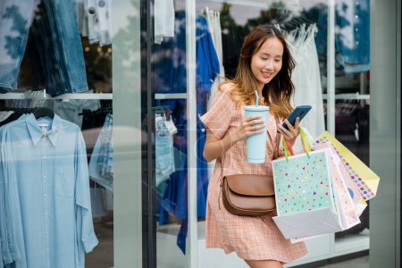 Téléchargez les photos : Mode de vie en ville : une femme navigue sur les applications shopping de son smartphone tout en tenant des sacs devant une fenêtre de magasin. - en image libre de droit