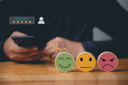 Photo for Mans using smartphone choosing a smiley face on a wooden block, evaluating customer experience. Surveying satisfaction with business services for excellent rating. - Royalty Free Image