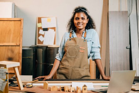 Photo for Portrait of american beautiful young black woman curly hair carpentry smiling standing and looking to camera on workspace table at workshop against wood or woodshop, National Carpenters Day - Royalty Free Image