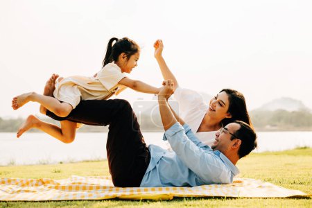 Foto de Un juguetón día en familia en el parque. El padre levanta a su hija en alto, mientras ella abre alegremente sus brazos y vuela como un avión, mientras que la madre mira con felicidad. Diversión familiar capturada en una fotografía - Imagen libre de derechos