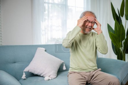 Foto de Dolor de cabeza. Hombre mayor asiático triste migraña crónica dolor masaje, anciano sostiene la cabeza con la mano para la enfermedad de dolor de cabeza en la sala de estar en el hogar, problemas de salud de la vejez, atención médica - Imagen libre de derechos